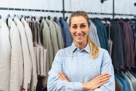 Confident tailor with arms crossed standing in front of suit jackets on hangers