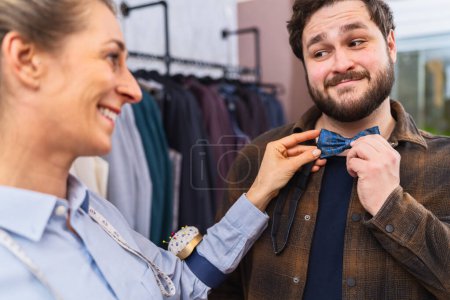 Tailor fitting a blue bow tie on a customer, both smiling, with clothing in background
