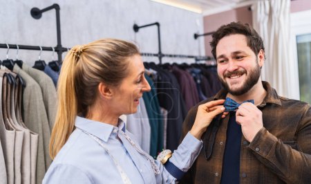 Saleswoman fitting a bow tie on a customer, both are smiling, racks of clothes behind in a men's clothing store
