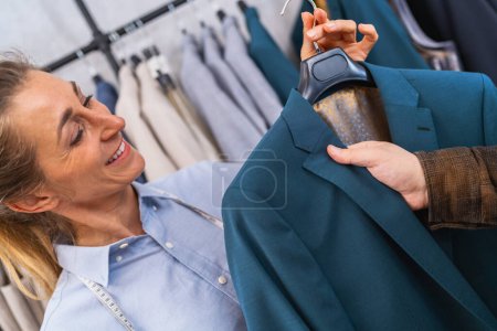 Smiling sailswoman showing a teal jacket to a male customer, clothes racks in the background at weeding store
