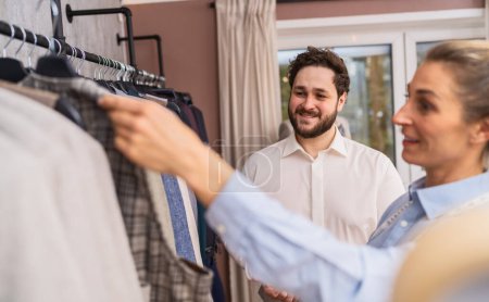 Schneiderin hält einem lächelnden männlichen Kunden in einem Hochzeitsgeschäft eine karierte Jacke hoch