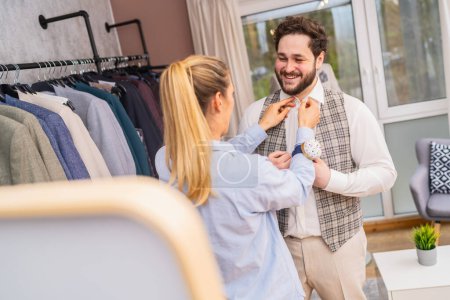 tailleur ajustant un gilet sur un homme souriant dans une boutique avec des étagères à vêtements
