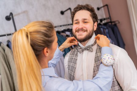 Tailor fitting a bow tie on a cheerful man in a fashion store with clothing racks