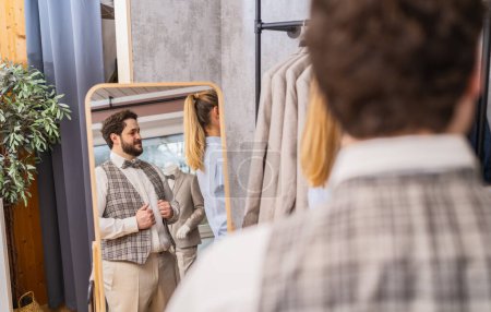 Man trying on a vest in front of a mirror in a clothing store with a tailor assisting