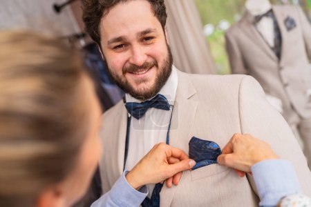 Man being fitted for a suit, tailor adjusts a blue pocket square, both smiling in a wedding store