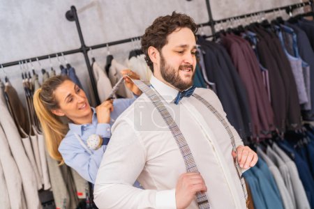 Tailor playfully pulling on client's suspenders, both smiling in a clothing store