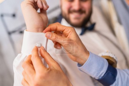 Tailor assists man with cufflinks, both hands visible, man smiles in background at a weddomg store