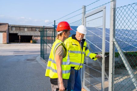 Foto de Dos ingenieros en chalecos de alta visibilidad inspeccionan un campo de paneles solares que entra en una puerta de la instalación - Imagen libre de derechos