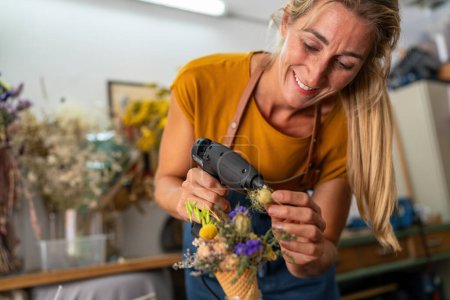 Téléchargez les photos : Fleuriste souriante utilisant un pistolet à colle sur un arrangement de cône de crème glacée florale - en image libre de droit