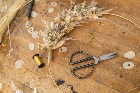 Photo for Dried flower arrangement and scissors on a wooden table with scattered petals and thread spool - Royalty Free Image