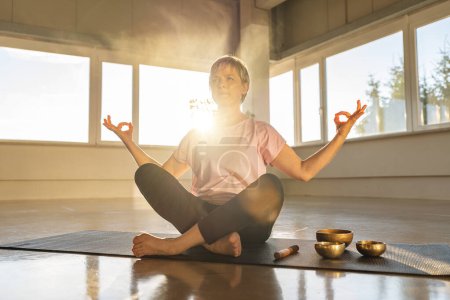 Frau in Meditationspose mit Klangschalen, heiterer Turnhallenatmosphäre, Sonnenaufgang