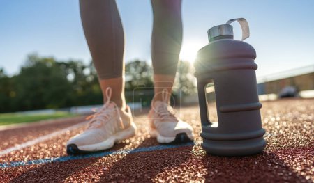 Foto de Primer plano de una botella de agua grande en una pista de atletismo con patas de mujer en el fondo - Imagen libre de derechos