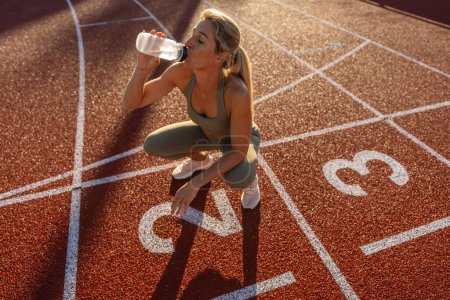 Foto de Atleta mujer beber agua mientras se agacha en un campo de pista al atardecer - Imagen libre de derechos