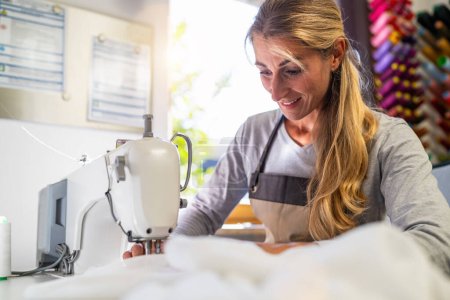 Foto de Mujer en un delantal está trabajando en una máquina de coser, costura de tela blanca en un espacio de trabajo bien iluminado con carretes de hilo de colores en el fondo - Imagen libre de derechos