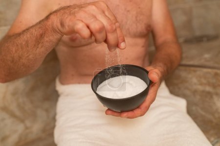 Älterer Mann hält im Dampfbad oder Hamam eine Schüssel Salz in der Hand, um die Haut für eine Körpermassage in einem Wellness-Resort zu peelen 