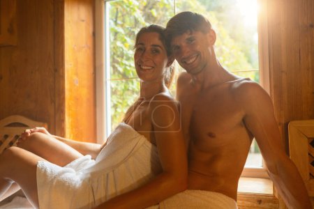 couple relaxes in a wooden finnish sauna, illuminated by soft sunlight filtering through a window. Both wear white towels, sharing a content and joyful moment. spa and wellness hotel or resort concept