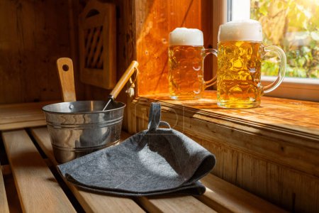 Zwei Bierkrüge mit schaumigem Bier ruhen auf einem Vorsprung und fangen das Sonnenlicht ein. In der Nähe stehen ein Metalleimer, eine Schöpfkelle und finnische Saunahüte auf einer Holzbank. Kur- und Wellness-Konzept Image