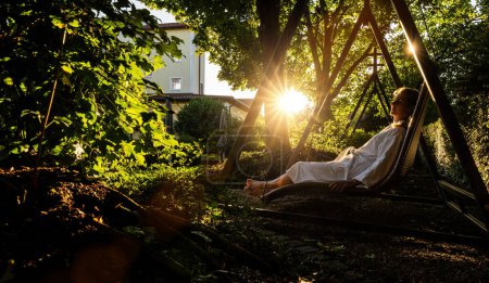 Frau im weißen Bademantel entspannt auf Weidenschaukel im Garten des Wellnesshotels