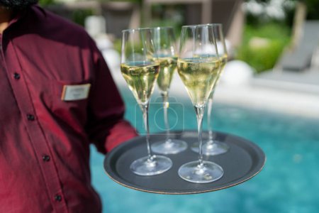 Waiter in a red shirt holding a tray with three glasses of champagne by a pool. Hotel travel concept image