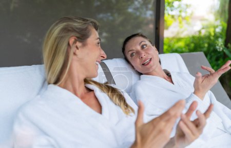 Two women in white bathrobes talking and gesturing on outdoor loungers at a spa wellness resort