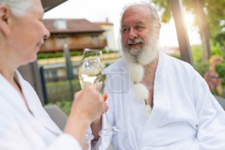 Seniorenpaar in weißen Bademänteln klimpert mit Sektgläsern, Mann schaut Frau im Kurhotel an