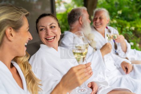Group of people in white bathrobes laughing and holding champagne glasses celebrating on a lounger at a spa wellness hotel