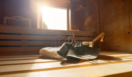 Sauna interior with felt hats, towels, ladle, and bucket, sunlight through window in a finnish sauna. Spa wellness hotel concept image.