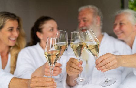 Gruppe von vier Personen in weißen Bademänteln prostet mit Champagnergläsern zu und feiert ein Ereignis in einem Wellness-Hotel