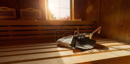 Wooden sauna with benches, window, towel, metal bucket, ladle, and felt hats in a finnish sauna. Spa wellness hotel concept image.