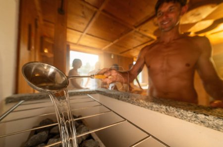 Close-up of man with ladle pouring water on hot sauna stones, with a blurred person in the background. Finish sauna spa wellness hotel concept image