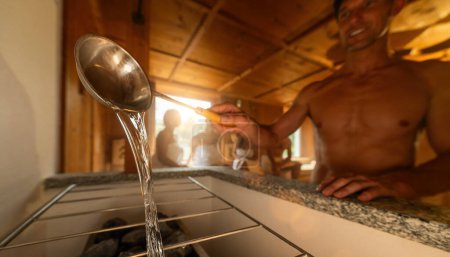Hombre vertiendo agua de un cucharón de sauna sobre rocas calientes en una estufa de sauna. Gente en el fondo. Terminar sauna spa wellness hotel concepto imagen.