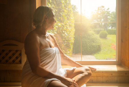 Frau sitzt auf einer Holzbank in einer Sauna und schaut aus dem Fenster