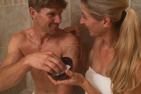 Nahaufnahme eines Paares in einem Dampfbad, das sich ein Salzpeeling teilt und einander anlächelt. Wellness spa hotel konzept image