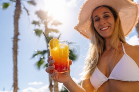 Frau im weißen Bikini und Strohhut bietet einen tropischen Cocktail an, sonniger Himmel mit Palmen. Party und Sommerurlaub Konzeptbild auf einer Karibik-Insel.