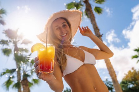 Smiling woman in white bikini and straw hat holding a tropical cocktail, palm trees, sunny sky at caribbean island