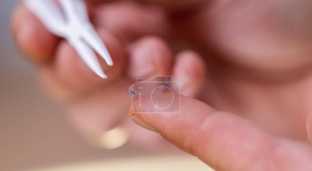 Close-up of a contact lens on a fingertip with a tweezer in the background.