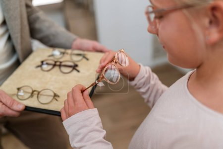 Mädchen beim Anprobieren einer neuen Brille. Sie betrachtet verschiedene Rahmen auf einem Tablett in einem Optikergeschäft.
