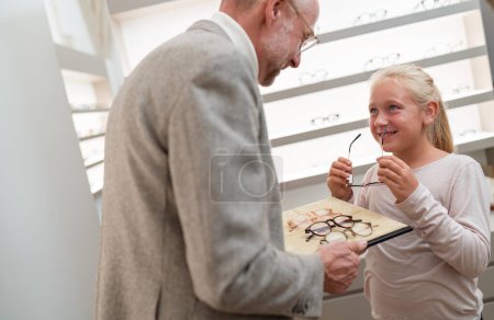 Smiling girl examining glasses with a optician assisting her by choosing eyeglasses in eyewear shop.