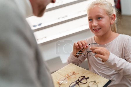 Girl holding glasses with optician and trying on new glasses. She is looking at eyewear selection in optician store. 