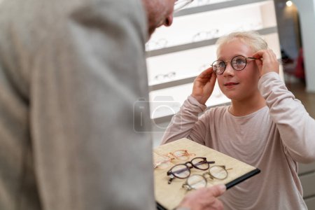 Child trying on round glasses. Optician holding eyewear tray in optical shop.