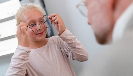 Junges Mädchen probiert neue Brille an und lächelt. Optiker untersucht, wie ein junges Mädchen in einem Brillengeschäft eine Brille anprobiert. 