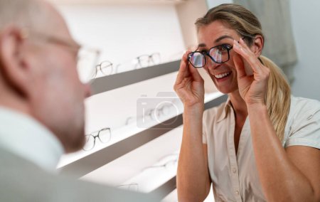 Smiling woman trying on black framed glasses. Optician in foreground. Eyewear shop interior