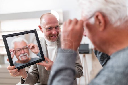 Optician holding a mirror for a older customer trying on glasses in a optician store