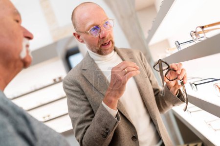 Foto de Optometrista presentando gafas graduadas al cliente en tienda óptica. Él está presentando la selección de gafas delante de la estantería de gafas - Imagen libre de derechos