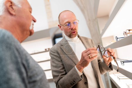 Optometrist presenting eyeglasses to customer in optical shop. Man is looking at eyewear selection in front of glasses shelf.