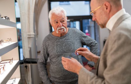 Foto de Hombre mayor escuchando al óptico sosteniendo un nuevo modelo de gafas en las manos en la tienda óptica - Imagen libre de derechos