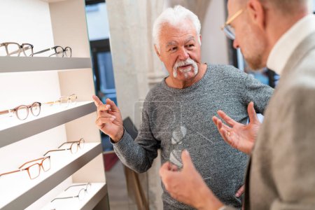 Foto de Hombre mayor haciendo gestos durante una conversación con un óptico en una tienda óptica con gafas expuestas en el fondo - Imagen libre de derechos