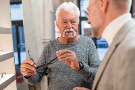 Foto de Hombre mayor probándose gafas nuevas y habla de ello con un óptico en la tienda - Imagen libre de derechos