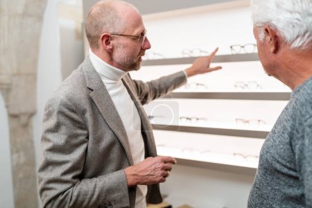 Foto de Óptico apuntando a diferentes gafas en exhibición al cliente senior en la tienda - Imagen libre de derechos