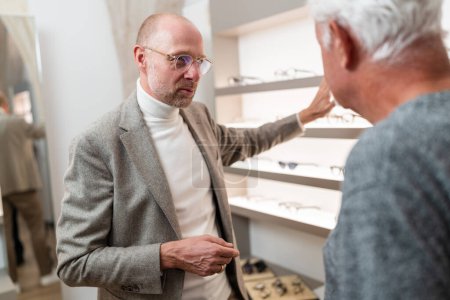 Optician showing eyewear to elderly client in optical store and explaining different eyewear models.